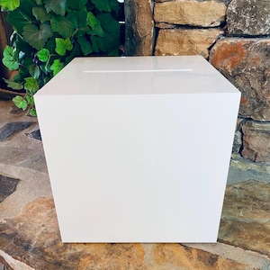 May include: A white square box with a slot on the top. The box is made of a smooth, glossy material and is sitting on a stone surface.