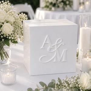 May include: A square white card box with a horizontal slot on top, featuring raised white lettering of the initials A & M on the front face. The box has a smooth, solid finish and is designed to hold cards for a special event or celebration.