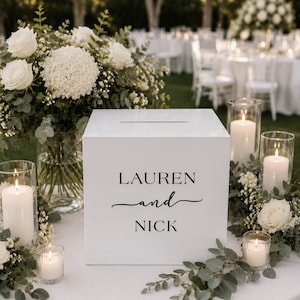 May include: A white square card box with a horizontal slot on top, featuring the black printed text LAUREN and NICK on the front face. The box is surrounded by white roses, green eucalyptus foliage, and several clear glass pillar candle holders.