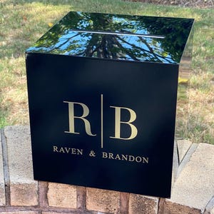 May include: Black acrylic wedding card box with a gold inscription that reads "R|B Raven & Brandon".