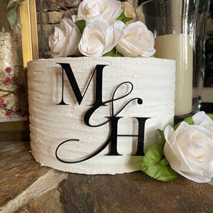 May include: A white cylindrical cake with a textured, horizontal-lined frosting finish. A black acrylic monogram topper featuring the letters M and H connected by an ampersand is attached to the front. White artificial roses decorate the top.