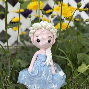 May include: A handmade crochet doll with a light pink face, white hair, and a blue dress. The doll's dress has a blue bodice and a sheer skirt with snowflake details. The doll is standing in a grassy area with yellow flowers in the background.