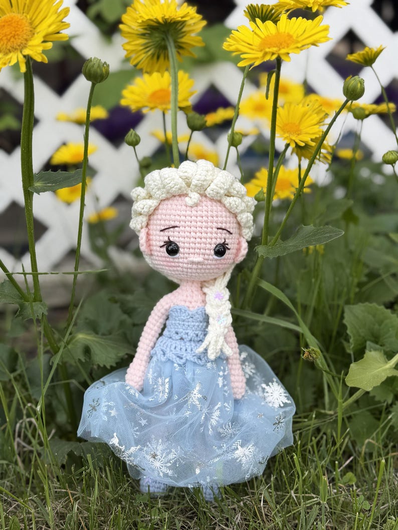 May include: A handmade crochet doll with a light pink face, white hair, and a blue dress. The doll's dress has a blue bodice and a sheer skirt with snowflake details. The doll is standing in a grassy area with yellow flowers in the background.