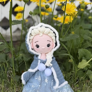 May include: A handmade crochet doll dressed in a blue and white outfit, reminiscent of a princess. The doll has a light pink face, black eyes, and a white braided hairstyle. The doll is wearing a blue dress with snowflake details and a blue hooded cape.