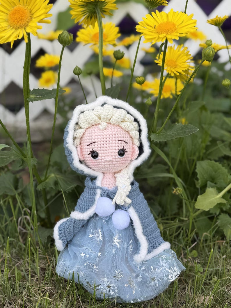 May include: A handmade crochet doll dressed in a blue and white outfit, reminiscent of a princess. The doll has a light pink face, black eyes, and a white braided hairstyle. The doll is wearing a blue dress with snowflake details and a blue hooded cape.