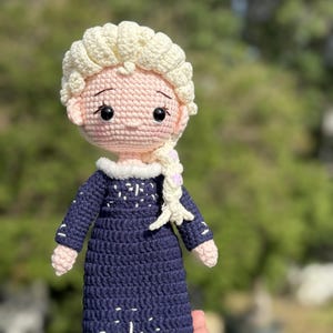 May include: A handmade crochet doll featuring a light skin tone, blonde hair in a braid, and a navy blue dress with white snowflake accents. The doll has a pink face and arms, and black eyes. The doll is approximately 8 inches tall.