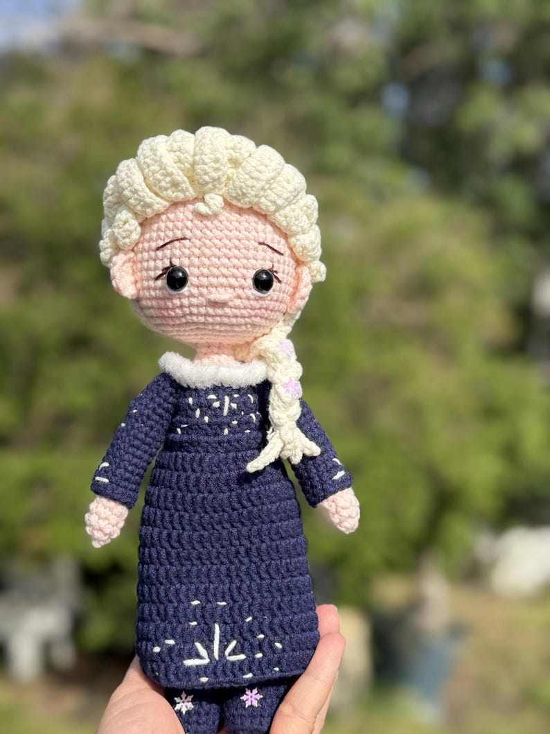 May include: A handmade crochet doll featuring a light skin tone, blonde hair in a braid, and a navy blue dress with white snowflake accents. The doll has a pink face and arms, and black eyes. The doll is approximately 8 inches tall.