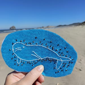 May include: A bright blue felt patch featuring a white embroidered outline of a seal. The design is embellished with small, scattered black beads that surround the seal, creating a textured effect against the solid blue fabric background.