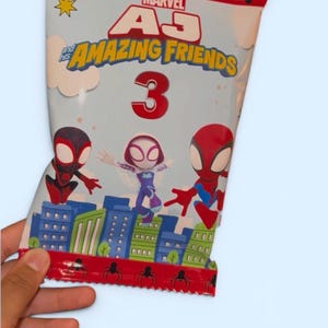 May include: A Marvel "Amazing Friends" snack bag featuring Spider-Man characters. The bag has a red top with spider graphics and the text "Marvel". The bag has a light blue background with the number "3" in red.