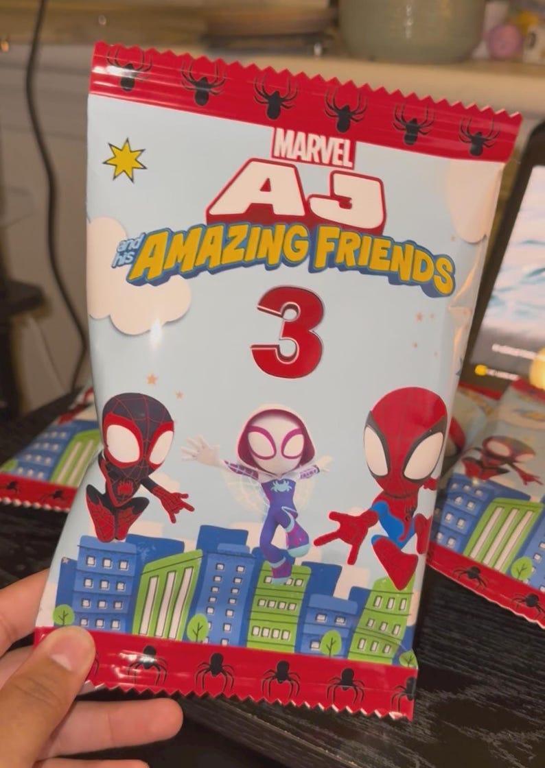 May include: A light blue snack bag featuring Marvel's "AJ and his Amazing Friends" with three Spider-Man characters. The bag has a red top with spider icons and the number 3. The background is light blue with city buildings.