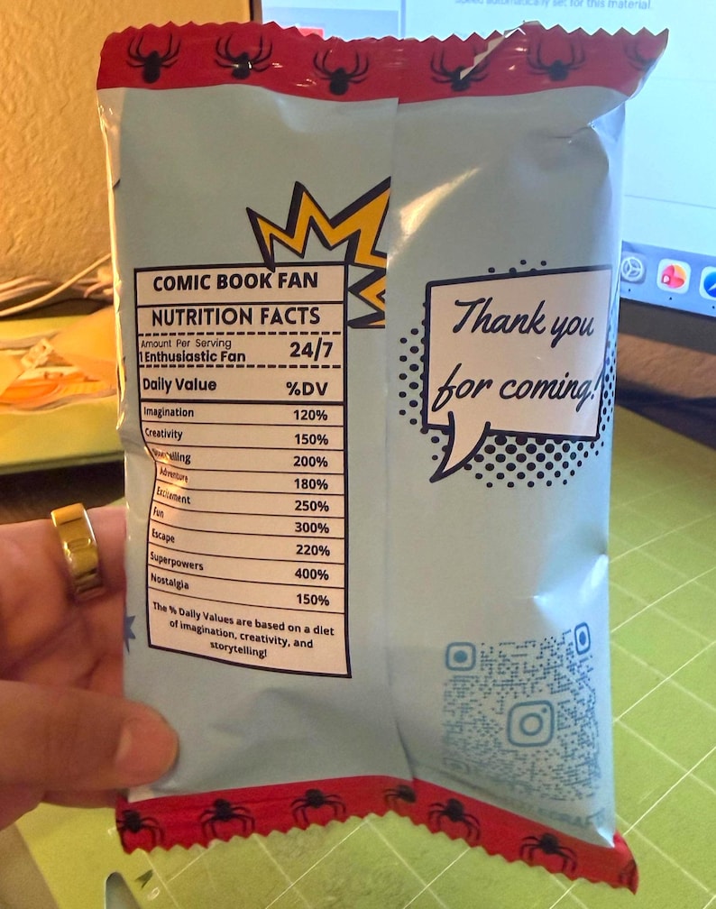 May include: A light blue snack bag with a red top featuring black spider graphics. The bag has text that reads "COMIC BOOK FAN" and "NUTRITION FACTS". The bag also has a speech bubble that says "Thank you for coming!"