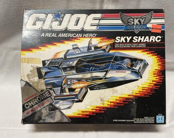 May include: A G.I. Joe Sky Patrol Sky Sharc toy box featuring a chrome-finished one-man attack craft with elevating twin cannons. The black box displays the vehicle in flight with orange exhaust flames, blue and white text, and a pilot figure.