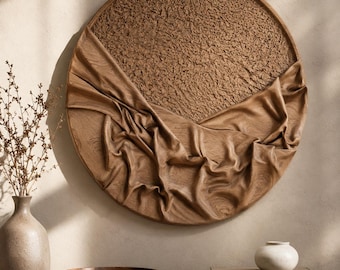 May include: A circular wall art piece featuring a textured upper half and a lower half with carved, draped fabric-like folds in a medium brown wood tone. The piece is mounted on a wall above a wooden table with a vase, bowl, and books.