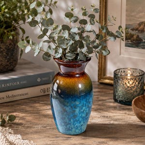 May include: A ceramic vase with a glossy finish that transitions from a dark brown rim to a vibrant blue base, filled with green eucalyptus stems. The vase has a rounded body and a flared neck, displaying a mottled, textured color gradient.