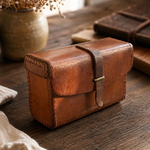 May include: A rectangular brown leather case with a flap closure secured by a vertical leather strap and a small brass buckle. The case features visible stitching along the edges and a weathered, textured surface with variations in the leather tone.