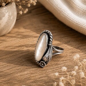 Può includere: Un anello di forma ovale con una pietra liscia color crema incastonata in una fascia di metallo color argento scuro ossidato. L'incastonatura è decorata con un bordo a corda intrecciata, motivi a foglia incisi e un piccolo fiore alla base.