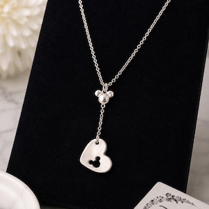 May include: A silver-toned necklace featuring a delicate chain, a small Mickey Mouse head charm, and a heart-shaped pendant with a Mickey Mouse head cutout. The necklace is accompanied by a white card with the word Disney printed in black script.