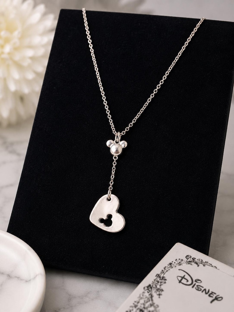 May include: A silver-toned necklace featuring a delicate chain, a small Mickey Mouse head charm, and a heart-shaped pendant with a Mickey Mouse head cutout. The necklace is accompanied by a white card with the word Disney printed in black script.