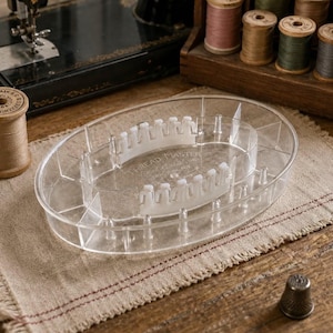 May include: A clear, oval-shaped plastic Thread Master organizer tray featuring multiple compartments, vertical pegs for thread spools, and two central white plastic racks with notched edges for securing thread ends, embossed with the text Thread Master.