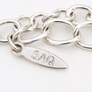 May include: A silver-toned chain necklace featuring a series of interconnected oval links. Attached to the chain is a small, elongated, leaf-shaped metal tag that is clearly engraved with the capital letters SAQ in a simple, sans-serif font.