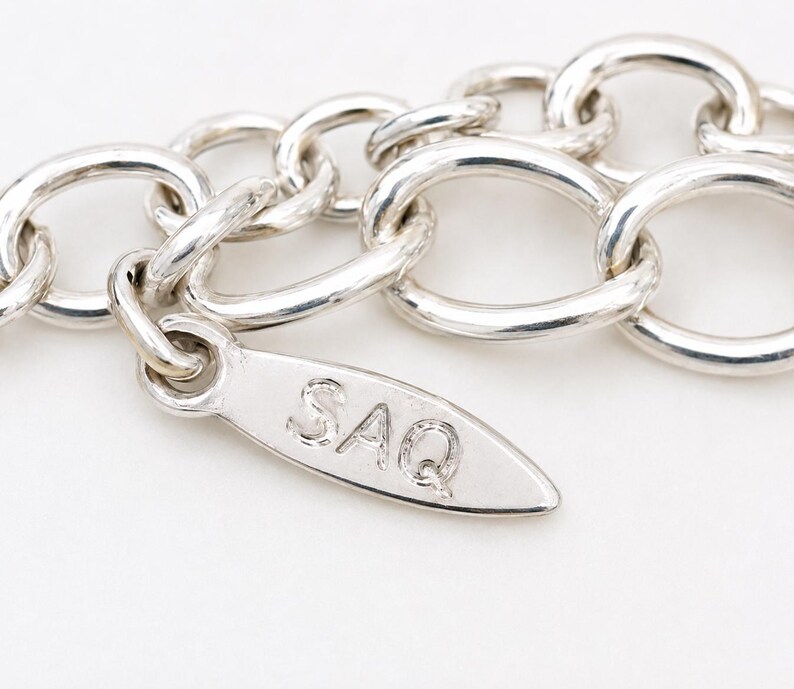 May include: A silver-toned chain necklace featuring a series of interconnected oval links. Attached to the chain is a small, elongated, leaf-shaped metal tag that is clearly engraved with the capital letters SAQ in a simple, sans-serif font.