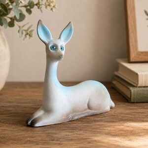 May include: A small, reclining deer figurine with a smooth, matte finish. The body is primarily white with soft, light blue shading on the ears, neck, and back. It features two prominent, sparkling blue inset eyes and dark gray hooves.