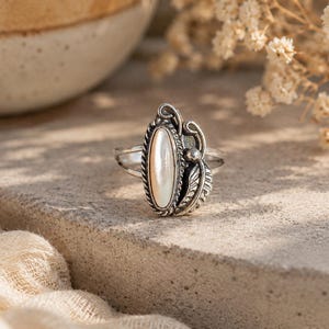 Pode incluir: Um anel de prata com uma pedra central ovalada e perolada. O anel apresenta detalhes decorativos de folhas e espirais. O anel está sobre uma superfície texturizada e de cor clara, com um fundo desfocado.