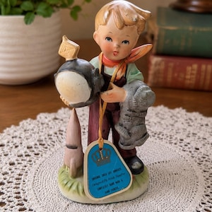 May include: A vintage ceramic figurine of a young boy holding a helmet and a gray object. The boy wears a green shirt, red scarf, and brown overalls. A blue tag with text hangs from the figurine. The base features a rocket and grass.