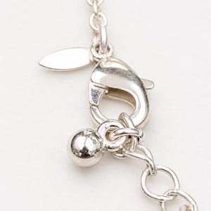 May include: A silver-toned metal necklace chain featuring a lobster claw clasp, a small smooth spherical charm, and a flat oval tag attached to the jump ring. The chain consists of round interlocking links with a polished, reflective finish.
