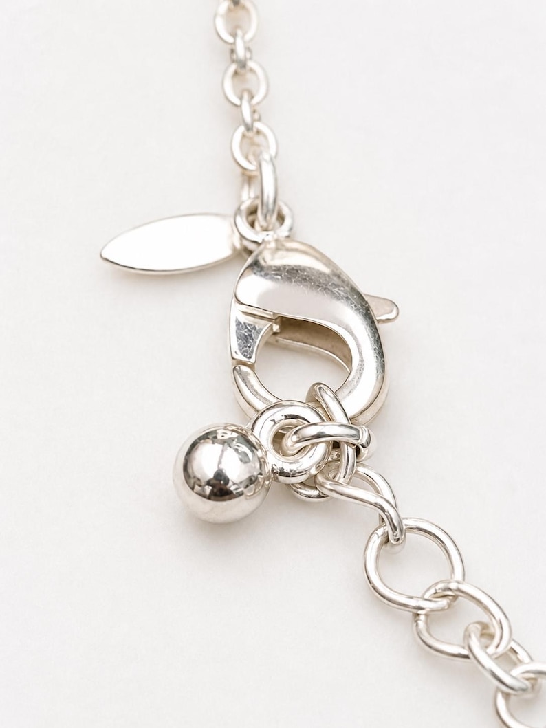 May include: A silver-toned metal necklace chain featuring a lobster claw clasp, a small smooth spherical charm, and a flat oval tag attached to the jump ring. The chain consists of round interlocking links with a polished, reflective finish.