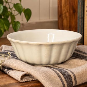 May include: A cream-colored ceramic bowl with a fluted, scalloped exterior design and a smooth, rounded rim. The bowl features a glossy finish and a deep, wide basin, resting on a folded beige and dark gray striped linen cloth.