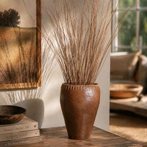 May include: A tall, brown vase filled with dried grasses sits on a wooden table. The vase has a textured surface and a stitched rim. A painting of a field and a stack of books are in the background.