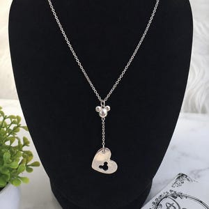 May include: A silver-toned necklace featuring a delicate chain with a small Mickey Mouse head charm and a heart-shaped pendant. The heart pendant contains a cutout in the shape of a Mickey Mouse head. The necklace is displayed on a black stand.