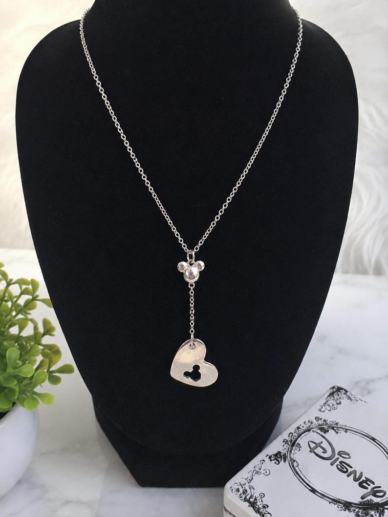 May include: A silver-toned necklace featuring a delicate chain with a small Mickey Mouse head charm and a heart-shaped pendant. The heart pendant contains a cutout in the shape of a Mickey Mouse head. The necklace is displayed on a black stand.