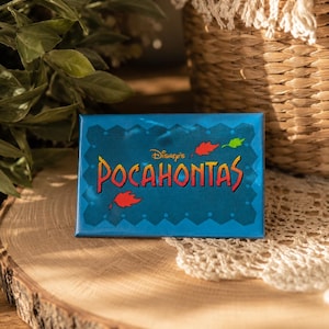 May include: A rectangular blue magnet featuring the text Disney's Pocahontas in red and yellow lettering. The design includes a scalloped border and small red and green leaf illustrations scattered around the central title on a dark blue background.