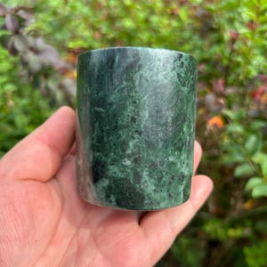 May include: A cylindrical vessel crafted from dark green marble, featuring natural veining and mottled patterns in shades of forest green, black, and white. The stone surface is polished to a smooth finish, showcasing the organic texture of the material.