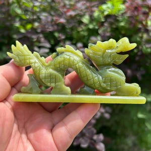 May include: A carved light green stone figurine of a dragon in a serpentine pose, featuring detailed scales, a horned head, and a flowing tail. The dragon is mounted on a flat, rectangular base of the same light green stone material.