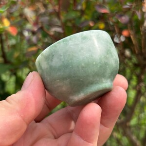 May include: A small, rounded bowl crafted from pale green stone with a smooth, polished surface. The bowl features subtle variations in color and texture throughout the material, with a wide, open top and a slightly tapered base.