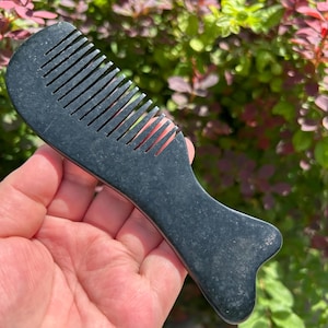 May include: A dark charcoal grey stone comb with a smooth, matte finish. The comb features a rounded top edge with evenly spaced teeth and a wider, ergonomic handle base that flares out slightly at the bottom, creating a solid, singular piece.