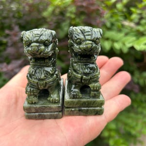 May include: Two small, dark green stone figurines carved in the likeness of guardian lions, each featuring detailed facial expressions, a mane, and a sturdy rectangular base. The sculptures are polished to a smooth, glossy finish with visible natural stone veining.