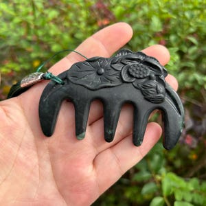 May include: A dark green, carved stone hair comb featuring a relief design of lotus flowers and leaves on the top edge. The comb has five wide, rounded teeth and is attached to a thin green cord with a small, textured silver-toned metal bead.