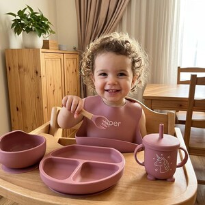 以下が含まれることがあります： ダスティローズ色のシリコン製食器セット。3つに仕切られたプレート、ボウル、蓋とストローと2つの持ち手が付いたユニコーン柄のカップ、木製ハンドルの小さなフォークが含まれており、すべて木製のトレイの上に配置されています。