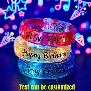 Könnte beinhalten: Drei gestapelte Armbänder aus durchscheinendem Kunststoff in Rot, Gelb und Blau, die Glitzer und Lichter enthalten. Das rote Armband trägt die Aufschrift GLOW PARTY, das gelbe Happy Birthday und das blaue Merry Christmas in schwarzer Schrift.