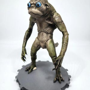 May include: A detailed figurine of a humanoid creature with green and tan skin, large blue eyes, and a frog-like appearance. The figure is posed on a gray, circular base with a gear-like edge.