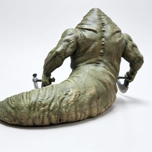 May include: A green, muscular, slug-like creature figurine with a textured, segmented back and a row of small bumps along the spine. The figure has two arms extended outward, each holding a small, dark-colored, curved blade-like weapon.