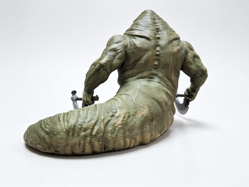 May include: A green, muscular, slug-like creature figurine with a textured, segmented back and a row of small bumps along the spine. The figure has two arms extended outward, each holding a small, dark-colored, curved blade-like weapon.