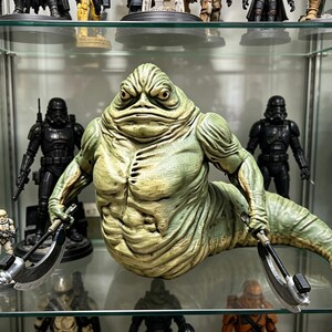 May include: A green, slug-like creature figurine with a textured, muscular torso and a long, tapering tail. The figure has a stern facial expression with deep-set eyes and holds a curved, metallic-looking blade in each of its two hands.