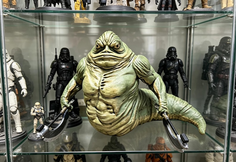 May include: A green, slug-like creature figurine with a textured, muscular torso and a long, tapering tail. The figure has a stern facial expression with deep-set eyes and holds a curved, metallic-looking blade in each of its two hands.