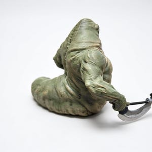 May include: A green, textured, slug-like creature figurine with a muscular upper body and a thick, segmented tail. The figure is depicted in a seated position, holding a silver-colored curved blade weapon in its right hand, with visible skin folds.
