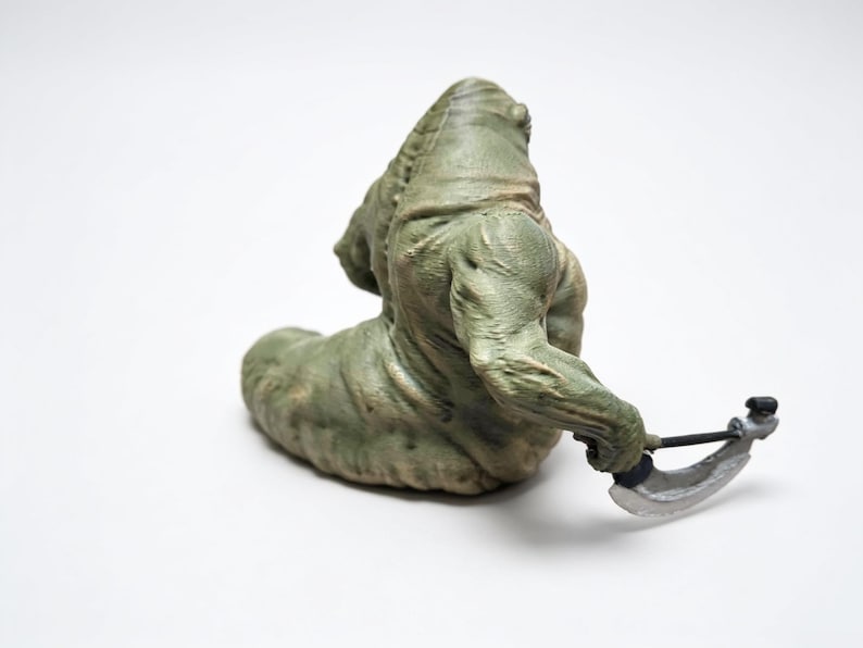 May include: A green, textured, slug-like creature figurine with a muscular upper body and a thick, segmented tail. The figure is depicted in a seated position, holding a silver-colored curved blade weapon in its right hand, with visible skin folds.